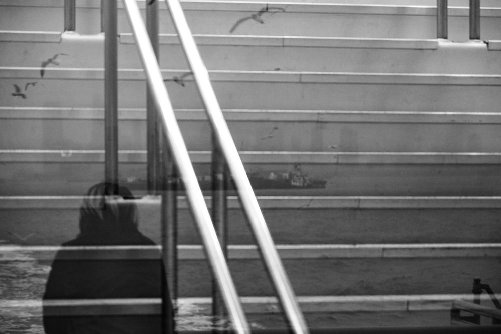 A black-and-white reflection shows a person on a stairway, overlaying an ocean view with a ferry and seagulls in Staten Island Ferry, Manhattan, New York.