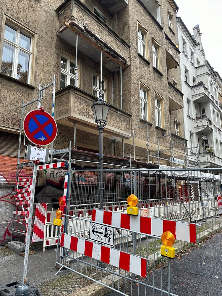 Ein Haus mit bröckelnden Balkonen, die mit einem Gerüst gestützt werden. Ein Gerüsttunnel für Fußgänger:innen. Dazu das für Berliner Verhältnisse belustigende Parkverbotsschild bis Ende 2023.