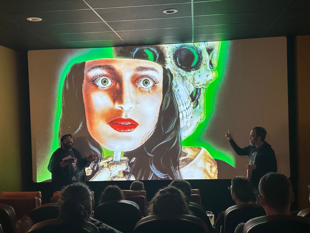 On the screen is the face of a green eyed girl with black hair and tears running down her face. Holding a stick with said Face ID a skull with a green glow. In the front presenting the show in theater four are Jenius and Greg.