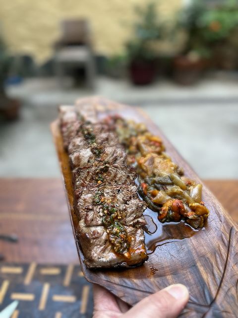 Thick slices of juicy Bife de Chorizo topped with chimichurri, served on a bed of charred bell peppers and onions. The meat glistens on a wooden platter, held up in a sunny courtyard, ready for the table.