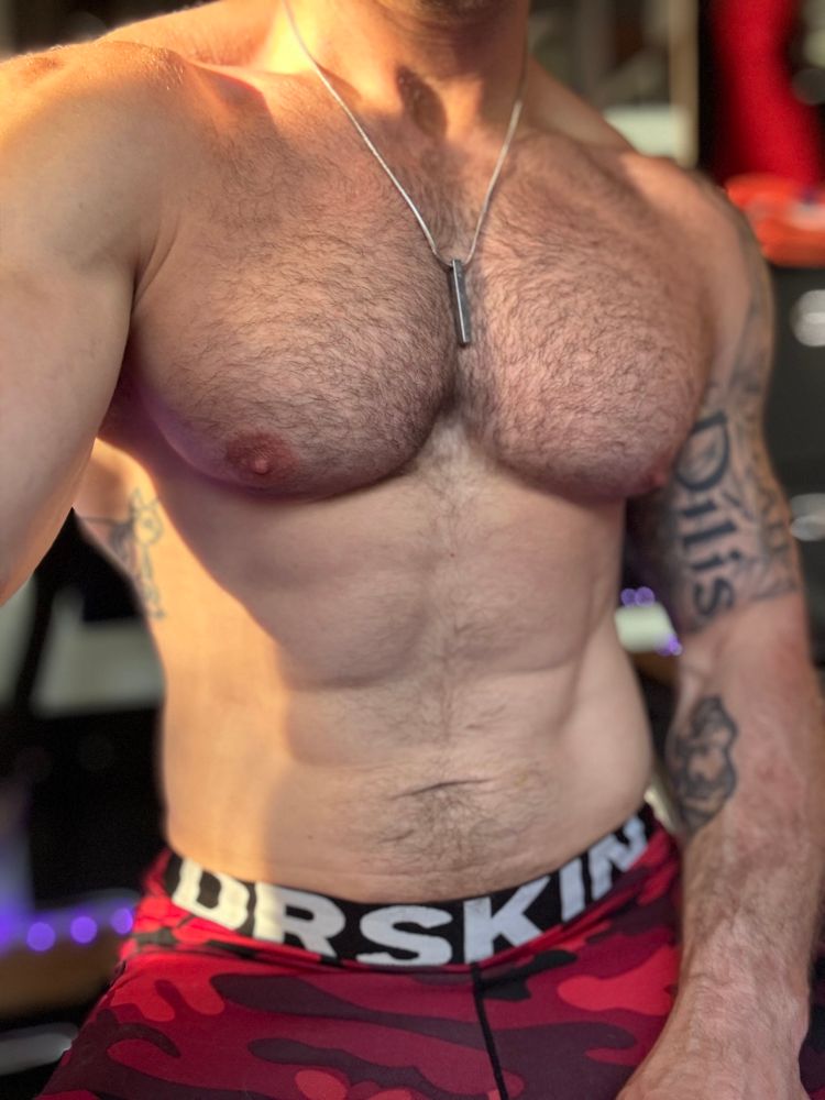 Shirtless gay guy torso with a silver chain around his neck and red and black camo tights that say Dr skin on the waistband 