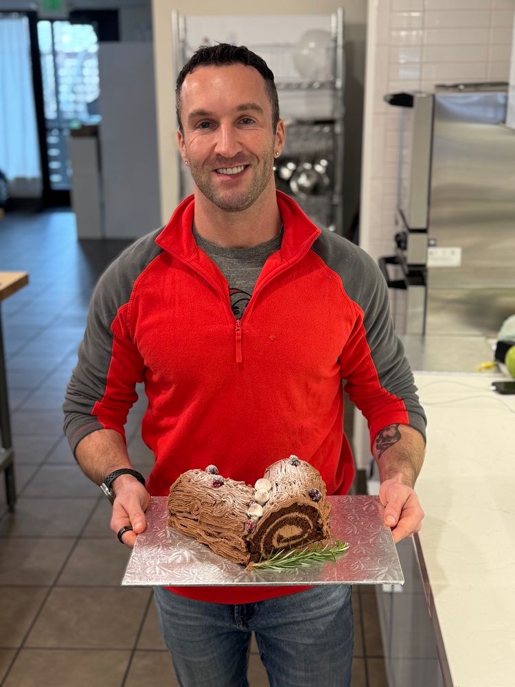 Gay guy in a red and grey fleece standing in a kitchen holding a silver platter with a chocolate yule log on it