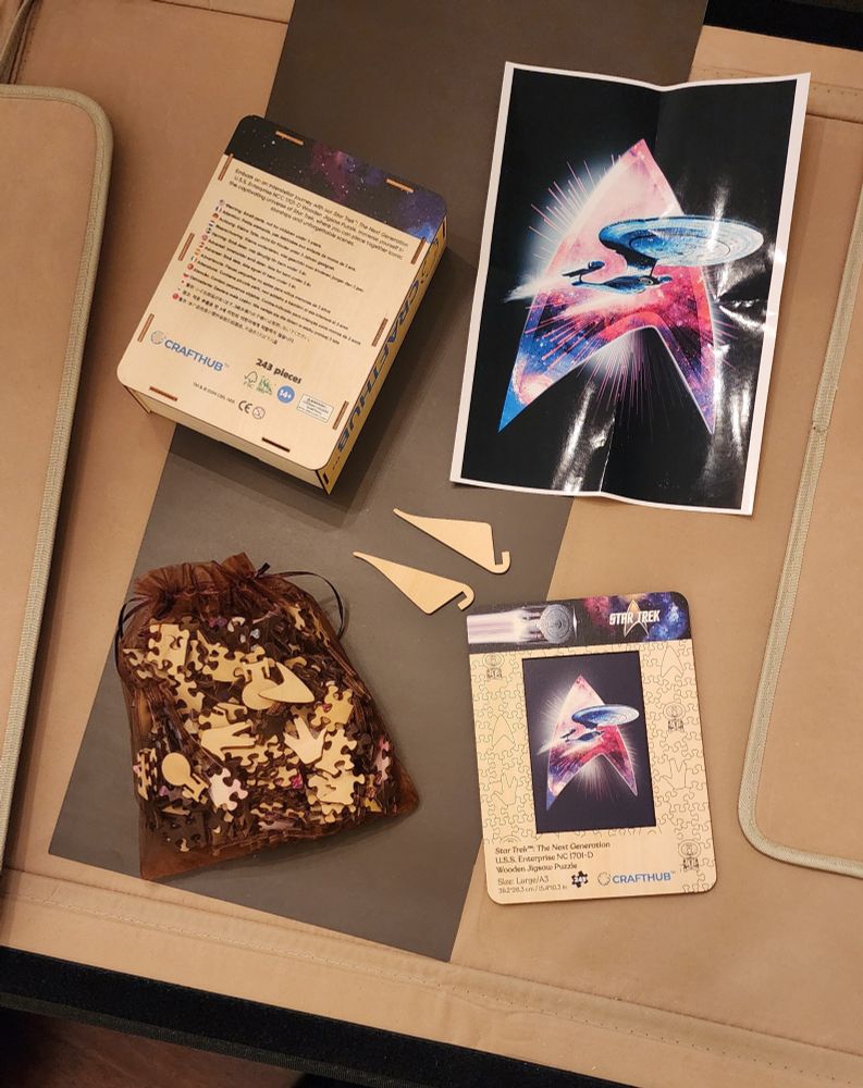 Wooden box containing Star Trek wooden puzzle is open amd contents displayed: bag of wooden puzzle pieces, wooden stands, and one sheet of puzzle design.