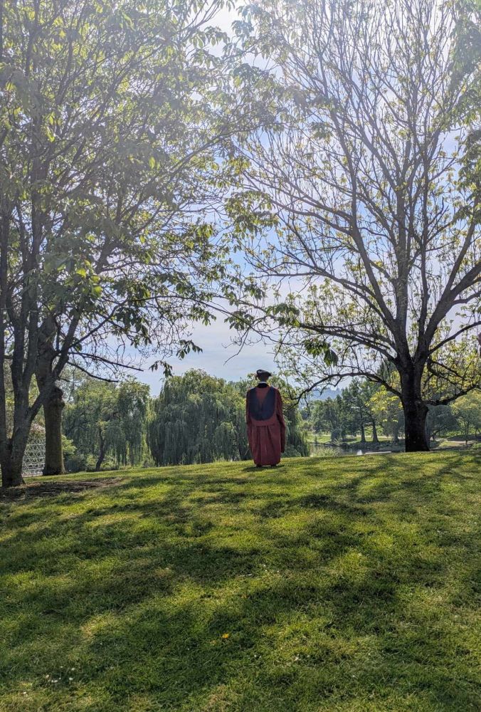 The summer skies are showing through the trees. A figure - me - standing in red robes looking away from the photographer and looking towards the lake. 