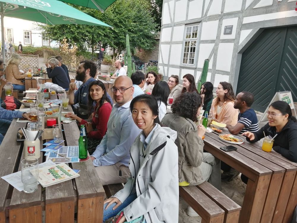 Researchers from across the Göttingen Campus attend a postdoc social meet-up. There are researchers from a range of disciplines - around 20 shown here - relaxing after a day's work in the lab or library or office. They sit outside at wooden benches.