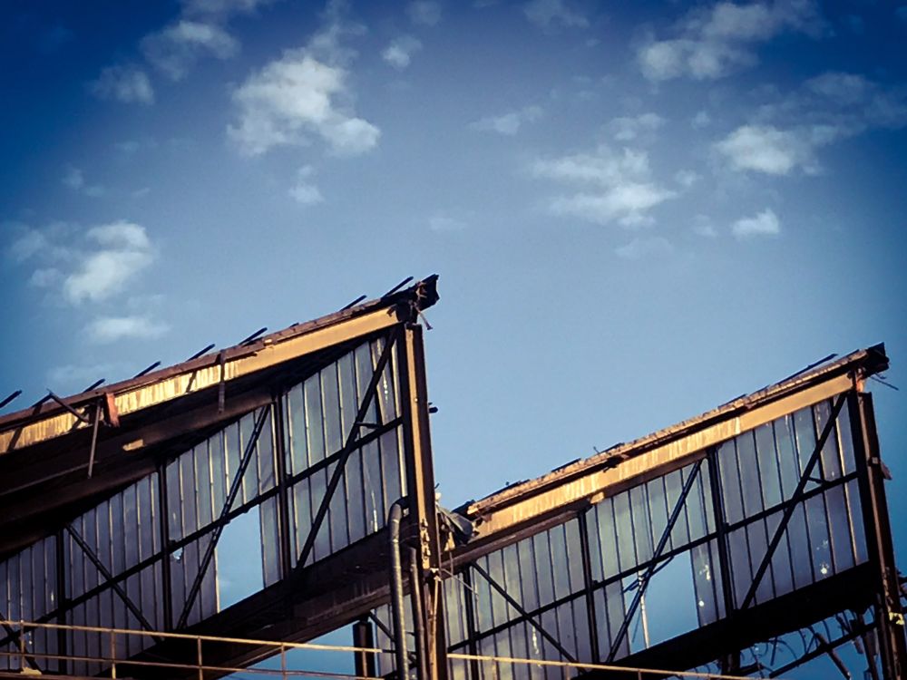 Image of an old factory roof made of glass panels (some broken) against the sky with some little fluffy clouds