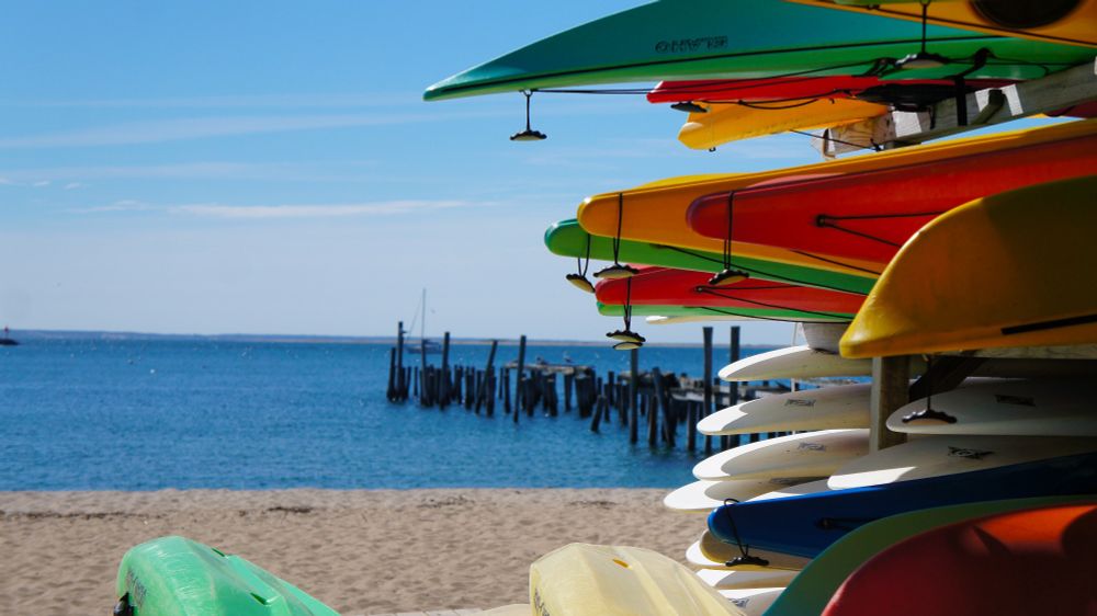 Image from the beach looking at a sandy beach with the sea and a broken pier in the background. In the foreground there are stacked canoes in multiple colours. 