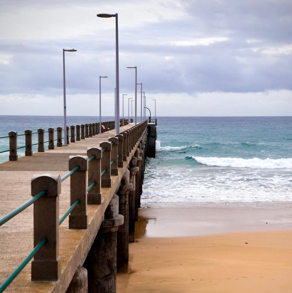 Picture of a concrete pier stretching across a sandy beach and into the sea. No one is on the pier. 