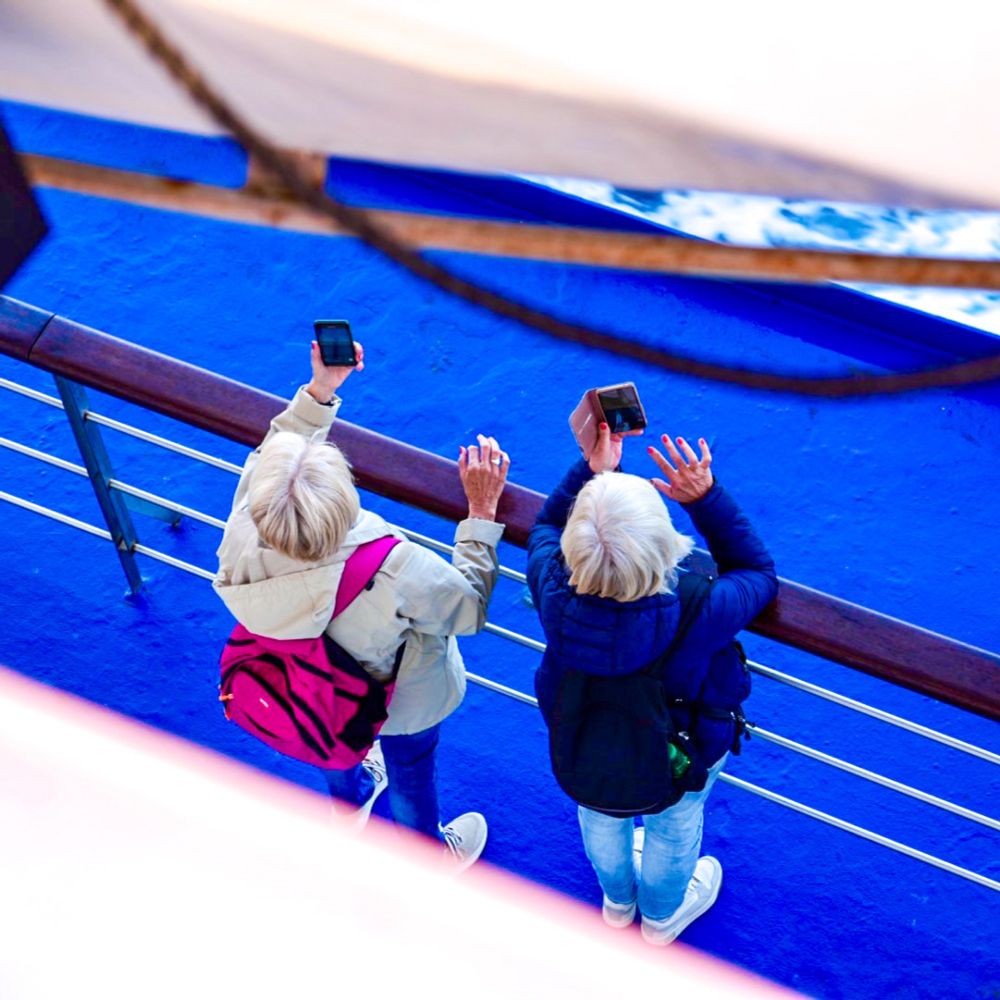Two silver haired women with their smartphone in their left hand held up while on a ferry. The picture is taken from above and the floor is bright blue. 
