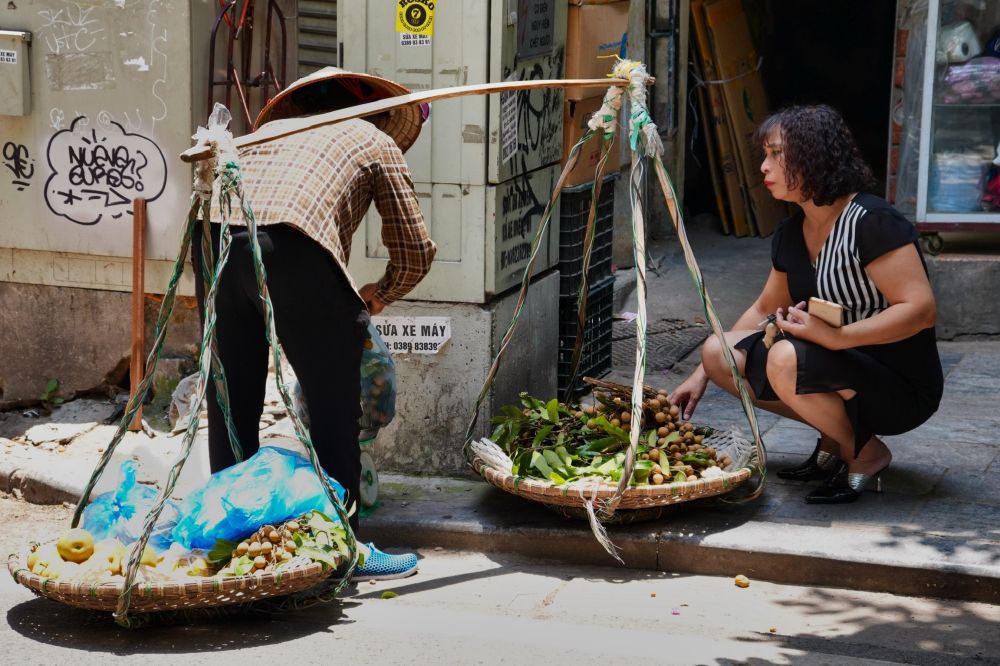 Traditional Vietnamese woman selling fruits from baskets hanging low from a bar of wood on her back. There is a younger woman kneeling down to look at her products. Taken on the side of the street in Hanoi