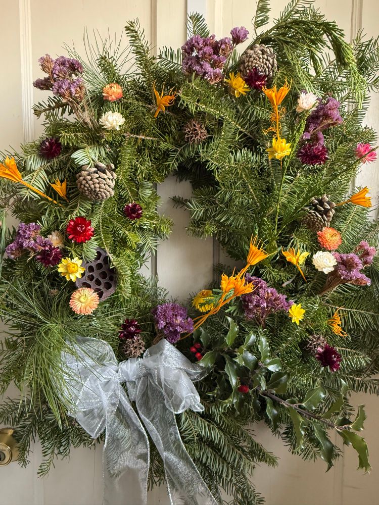 A large (for the size of the door at least) wreath with an evergreen backdrop with colorful dried flowers and seed heads mostly in purple, orange, yellow, and white; three pine cones and an elaborate brown honeycomb-like seed head; a sprig of holly with some red berries; some other sprigs from evergreens; and a shimmery opaque silver-white bow