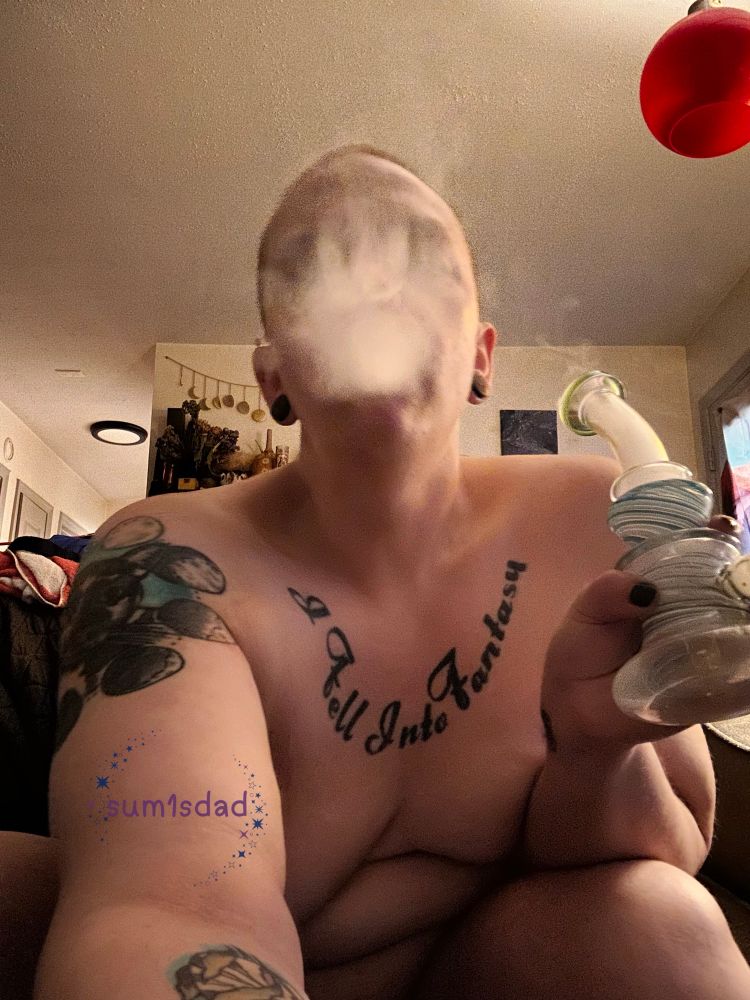 Larzipan sits naked on the couch, their breasts concealed by their arms- one extended holding the camera, the other holding their smoking apparatus. Smoke is billowing from their mouth, obscuring their face. 