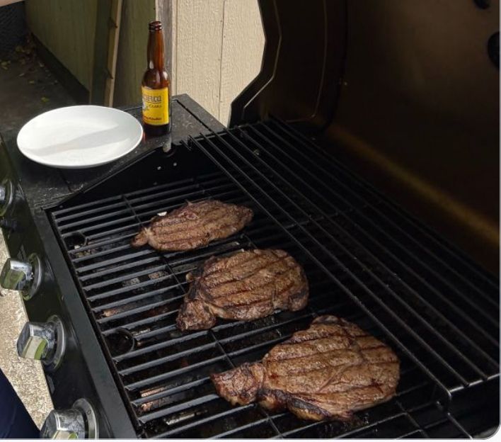 Photo of a grill with 3 steaks cooked with grill marks. A plate and beverage in a glass bottle are in the background.