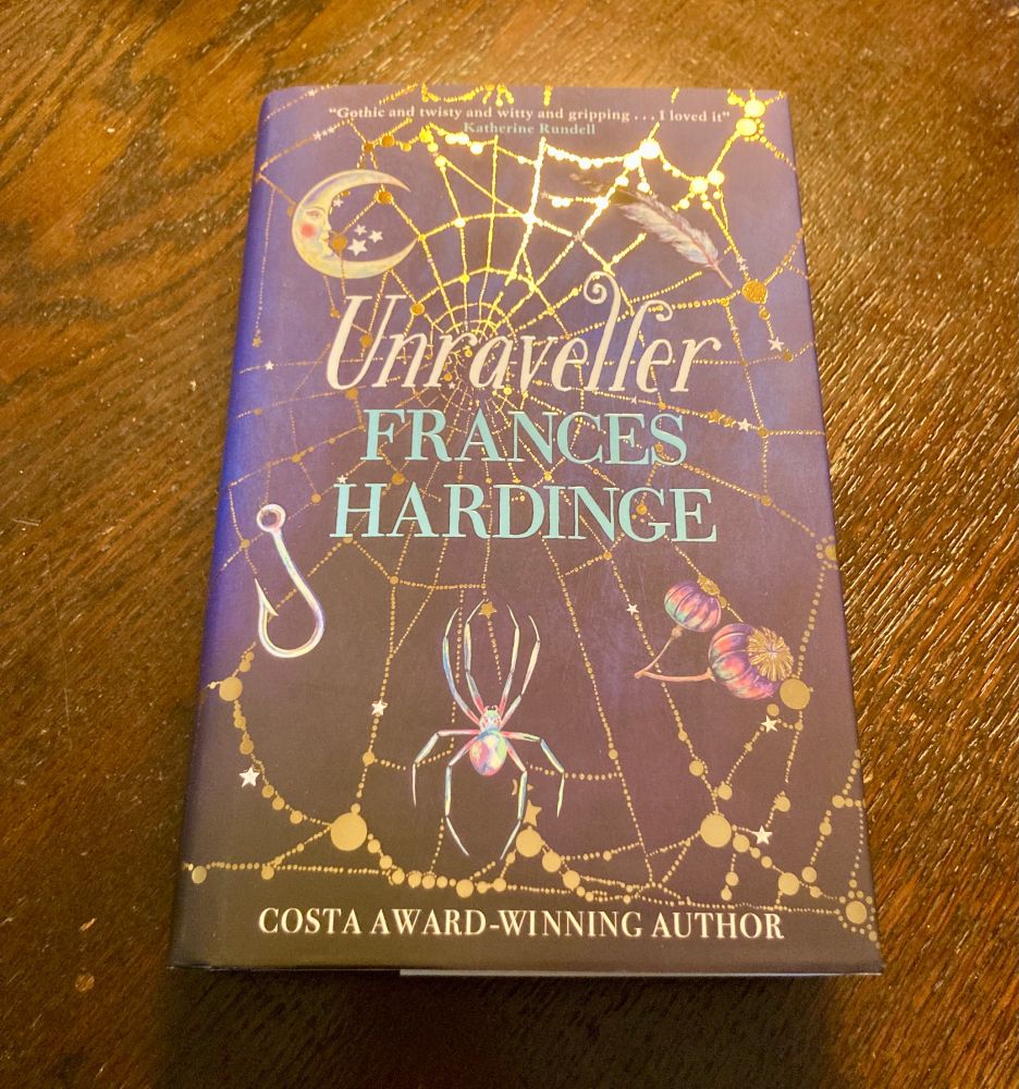 The jacket of "The Unraveller" by Frances Hardinge. Arranged around title and author's name are 5 key objects from the story: the moon, a feather, seed pods, a spider, and a fish hook. A spider web printed in metallic copper appears behind them, over a dark purple/black background.