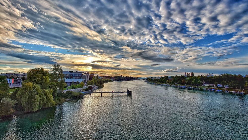 Rheinkilometer 1 in Konstanz. In der Mitte ist der Rhein. Darin steht eine Schiffsanlegestelle, links davon am Ufer ein Mehrzweckgebäude. Rechts sind Boote am Ufer festgemacht. An beiden Seiten des Ufer stehen Gebäude. Am Horizont ist der Bodensee zu sehen sowie zwei weitere Brücken.