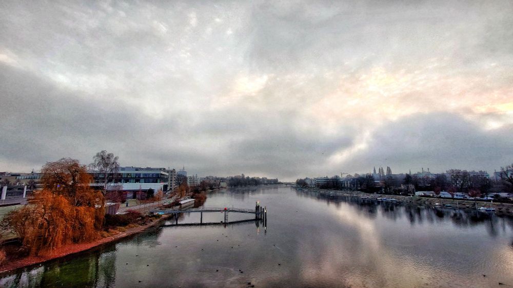 Rheinkilometer 1 in Konstanz. In der Mitte ist der Rhein. Darin steht eine Schiffsanlegestelle, links davon am Ufer ein Mehrzweckgebäude. Rechts sind Boote am Ufer festgemacht. An beiden Seiten des Ufer stehen Gebäude. Am Horizont ist der Bodensee zu sehen sowie zwei weitere Brücken.