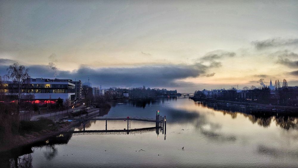 Rheinkilometer 1 in Konstanz. In der Mitte ist der Rhein. Darin steht eine Schiffsanlegestelle, links davon am Ufer ein Mehrzweckgebäude. Rechts sind Boote am Ufer festgemacht. An beiden Seiten des Ufer stehen Gebäude. Am Horizont ist der Bodensee zu sehen sowie zwei weitere Brücken.