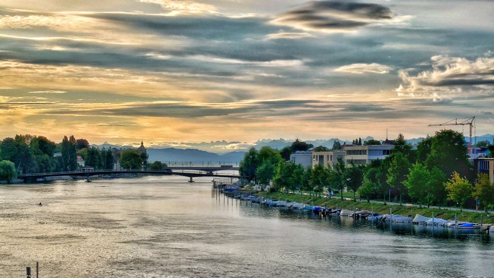 Der Seerhein in Konstanz wird von zwei Brücken überspannt. Am Horizont - über dem Bodensee - sind leicht die Alpen zu erkennen. Am rechten Rand des Bildes sieht man die Gebäude der HTWG, am Ufer sind Boote festgemacht.