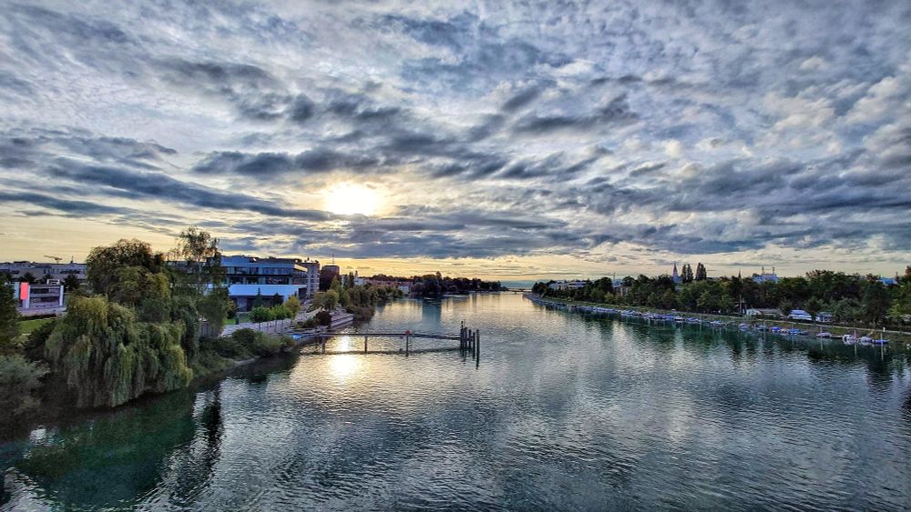 Rheinkilometer 1 in Konstanz. In der Mitte ist der Rhein. Darin steht eine Schiffsanlegestelle, links davon am Ufer ein Mehrzweckgebäude. Rechts sind Boote am Ufer festgemacht. An beiden Seiten des Ufer stehen Gebäude. Am Horizont ist der Bodensee zu sehen sowie zwei weitere Brücken.