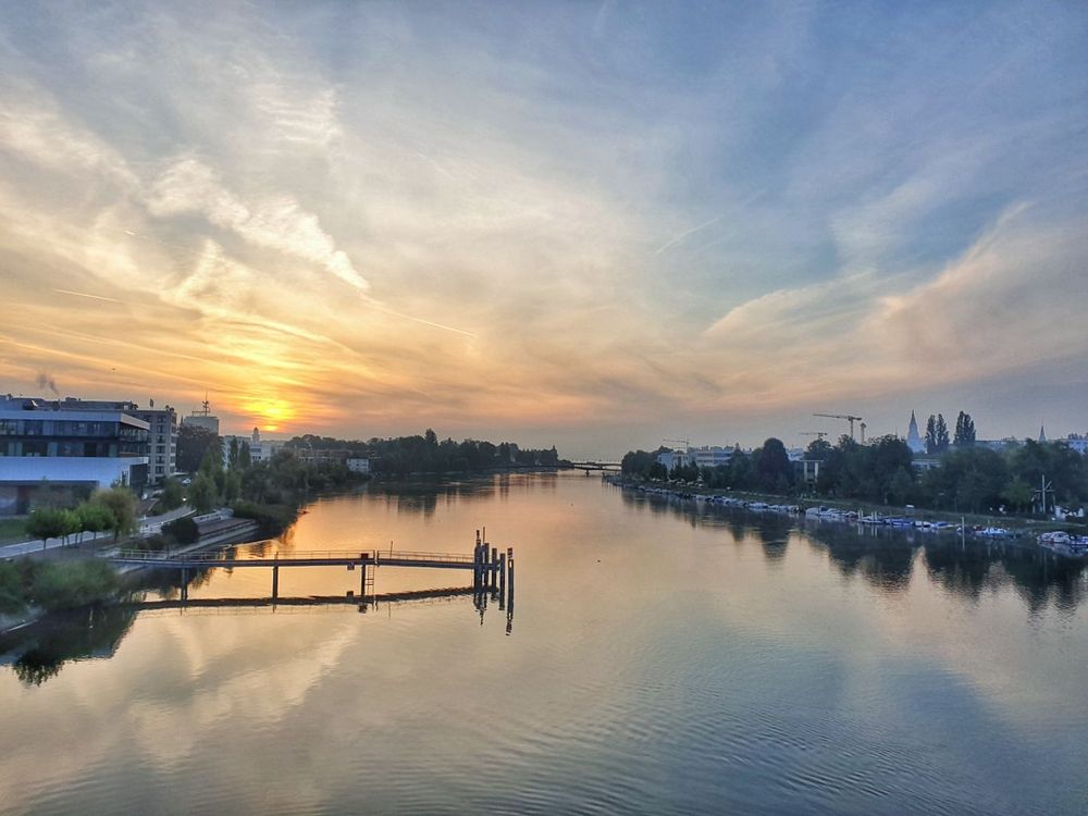 Rheinkilometer 1 in Konstanz. In der Mitte ist der Rhein. Darin steht eine Schiffsanlegestelle, links davon am Ufer ein Mehrzweckgebäude. Rechts sind Boote am Ufer festgemacht. An beiden Seiten des Ufer stehen Gebäude. Am Horizont ist der Bodensee zu sehen sowie zwei weitere Brücken.