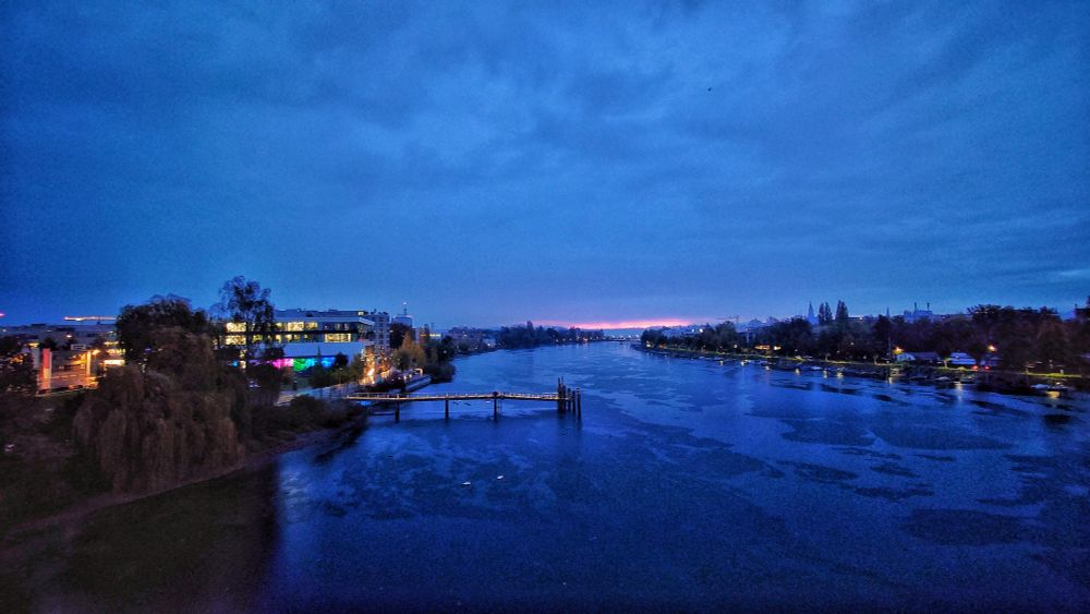 Rheinkilometer 1 in Konstanz. In der Mitte ist der Rhein. Darin steht eine Schiffsanlegestelle, links davon am Ufer ein Mehrzweckgebäude. Rechts sind Boote am Ufer festgemacht. An beiden Seiten des Ufer stehen Gebäude. Am Horizont ist der Bodensee zu sehen sowie zwei weitere Brücken.