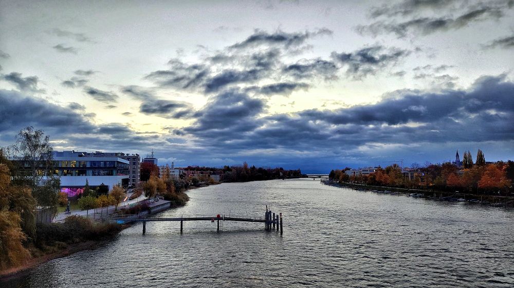 Rheinkilometer 1 in Konstanz. In der Mitte ist der Rhein. Darin steht eine Schiffsanlegestelle, links davon am Ufer ein Mehrzweckgebäude. Rechts sind Boote am Ufer festgemacht. An beiden Seiten des Ufer stehen Gebäude. Am Horizont ist der Bodensee zu sehen sowie zwei weitere Brücken.