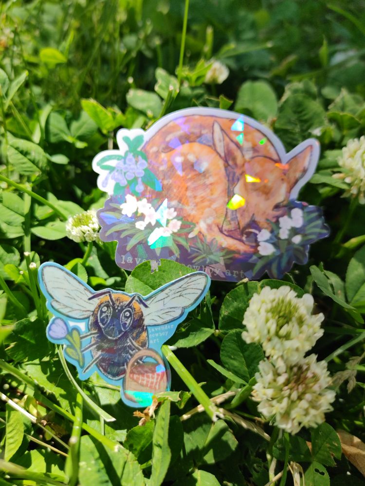 A second photo with the same subject matter as the previous, but now the deer sticker is in the background and the holographic bee is focused and in front. The bee sticker is an stylized illustration of a bumble bee flying towards the viewer, holding a purple tulip with one set of legs and a wicker picnic basket with another set. 