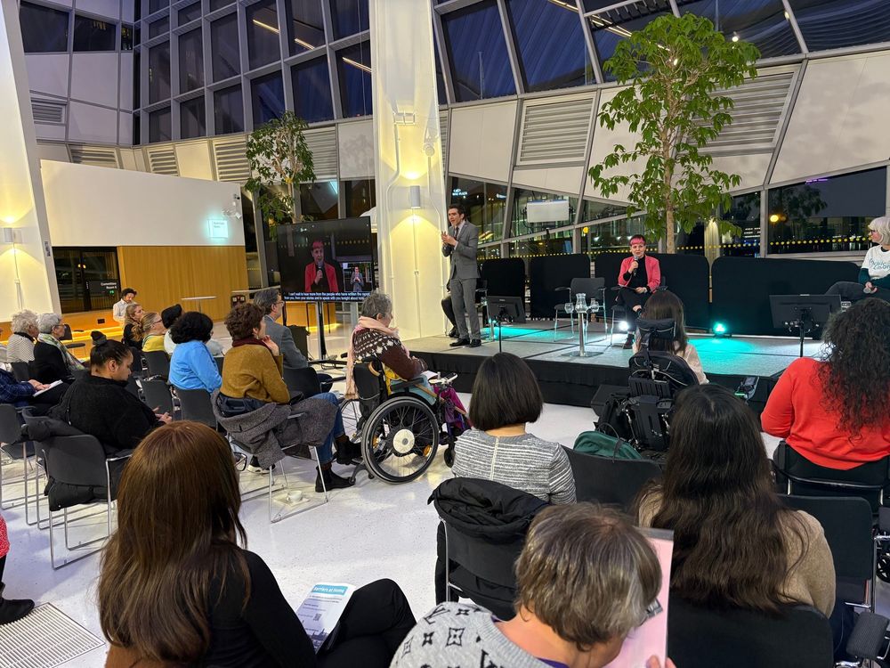 London Living Room at City hall full of people watching the stage. Zoë is in a pink jacket on stage speaking into a microphone. Someone signing next to Zoë