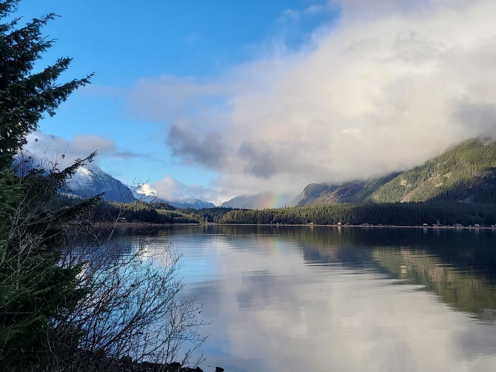 A view of Lutak Inlet in Haines Alaska on a bright November morning with some puffy clouds over the mountains on the right and a rainbow reflected in the water 