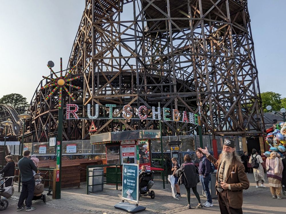 A grey-bearded man wearing a tweed jacket and a tweed fez by Fez-O-Rama motions appreciatively at a wooden rollercoaster named the "Rutschebanen" that was built in 1932.