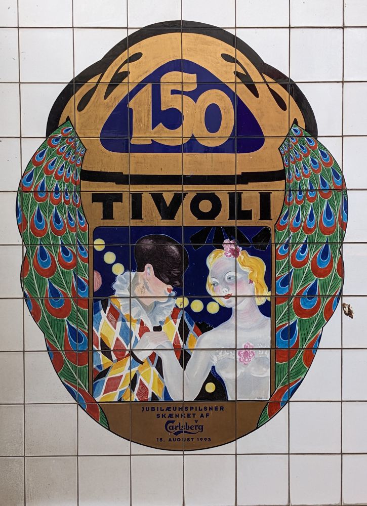 A tile mural commemorating 150 years of the  operation of Tivoli Gardens, the third-oldest operating amusement park in the world. The painted ceramic tiles depict stylized peacock feathers flanking a scene of a harlequin holding the hand of a ballerina.