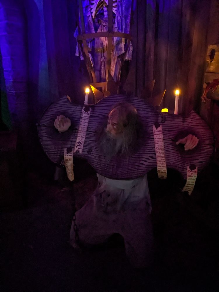A sad, pathetic wretch with scraggly long hair and a grey beard who wears filthy, tattered rags kneels in darkness while wearing a detached pillory that is festooned with bells, a crown of rusty iron spikes, strips of parchment with calligraphic writing and sigils, and has three illuminated candles mounted to the top of the antiquated wood frame.