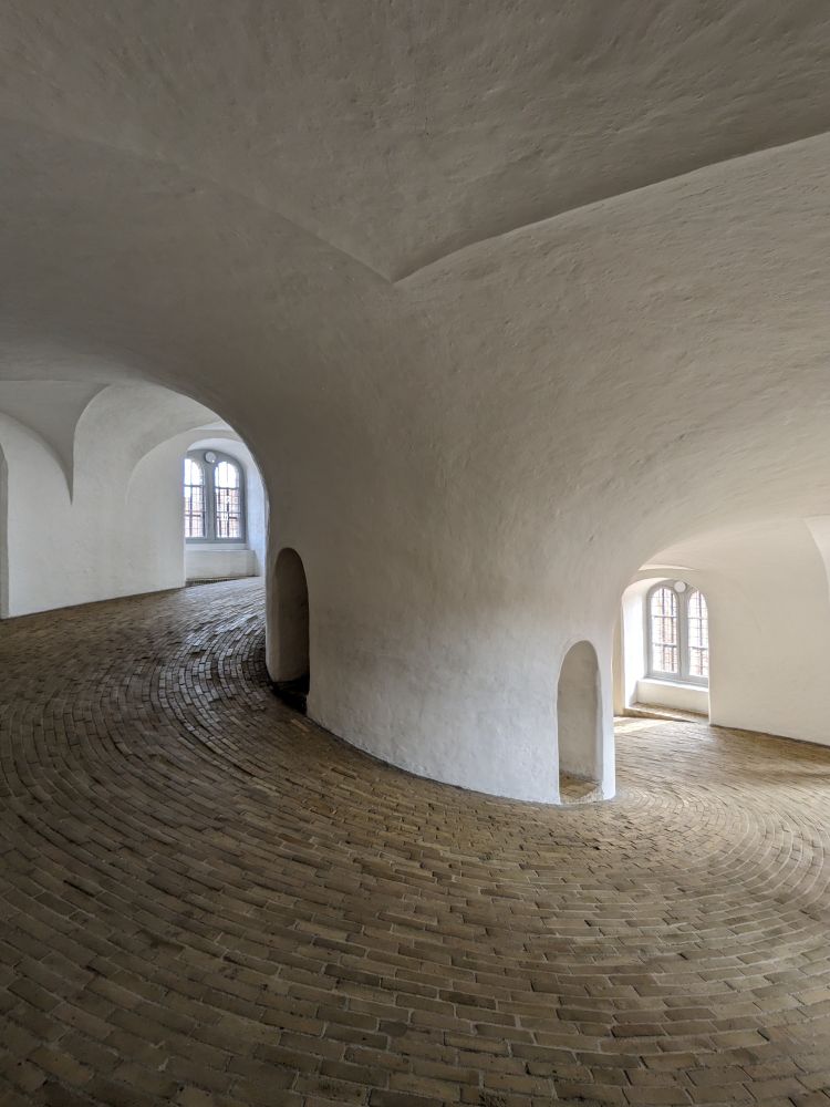 The interior of the Rundetaarn in Copenhagen, a cylindrical brick tower constructed in 1642. The walls are smooth white plaster transitioning into arches overhead, and the floor is a helical composition of tightly packed tan bricks. There are arched niches within the central spine of the structure.