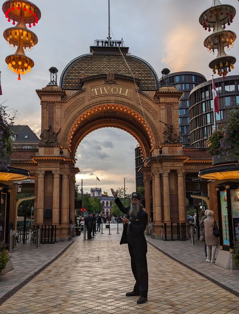 A grey-bearded man in a suit stands inside Tivoli Gardens at dusk, motioning appreciatively at the Baroque Revival gateway to Tivoli Gardens, an amusement park in Copenhagen, Denmark.