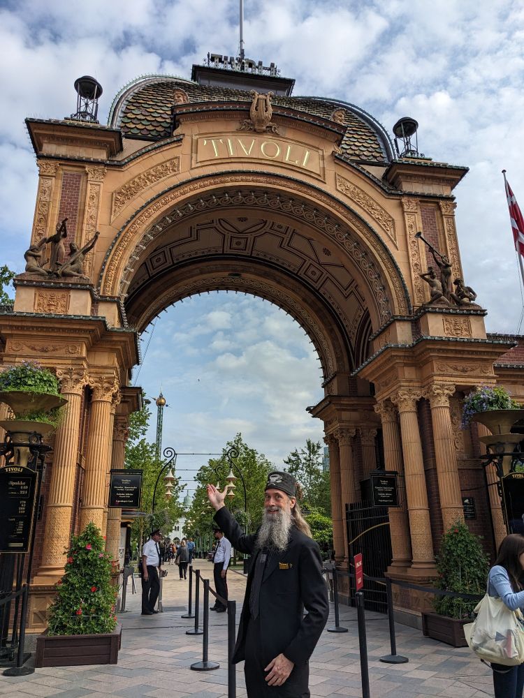A grey-bearded man in a suit stands outside a baroque revival gateway to Tivoli Gardens, an amusement park in Copenhagen, Denmark.