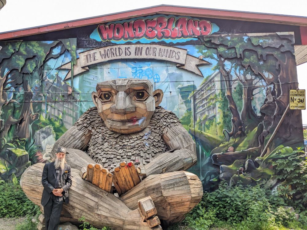A grey-bearded man in a black suit and wearing a grey tweed fez handcrafted by Fez-O-Rama leans up against a wooden troll statue sculpted by Thomas Dambo located in Freetown Christiania within Copenhagen, Denmark.