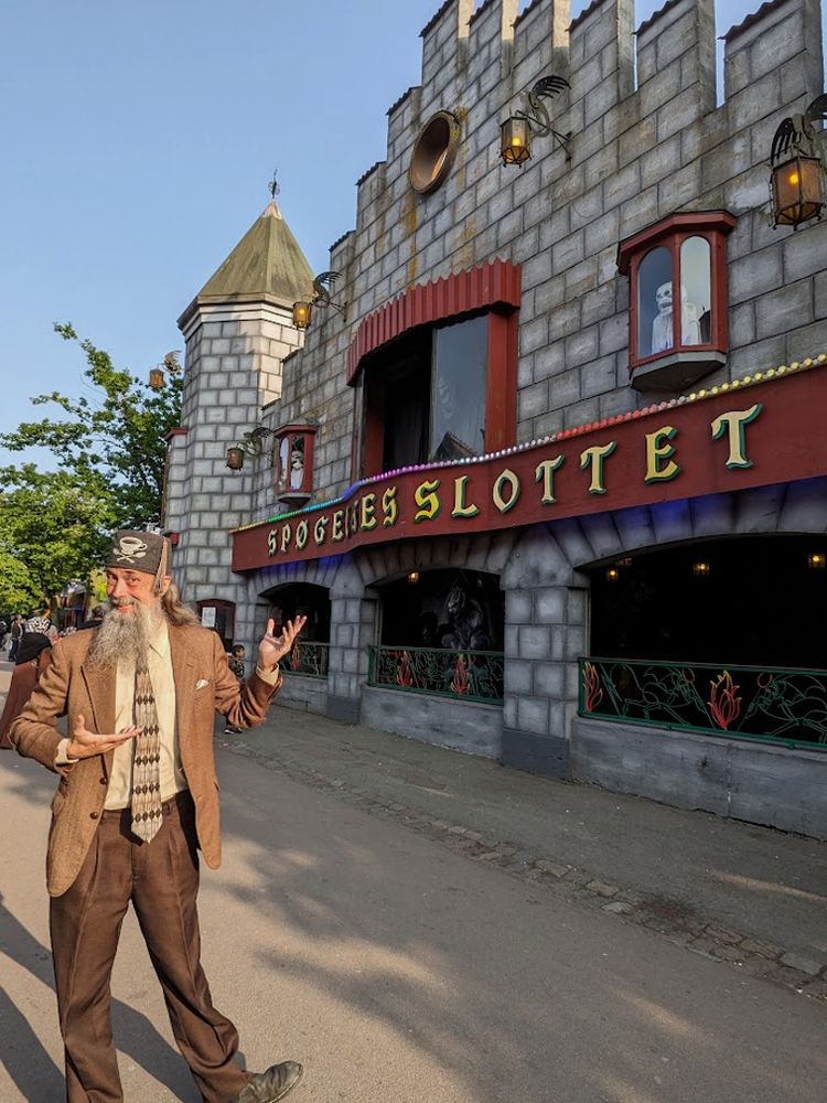 A grey-bearded man wearing a tweed jacket and a tweed fez by Fez-O-Rama happily motions at an amusement park Dark Ride called "Spøgelses Slottet," located at Bakken, the world's oldest amusement park located in Denmark.
