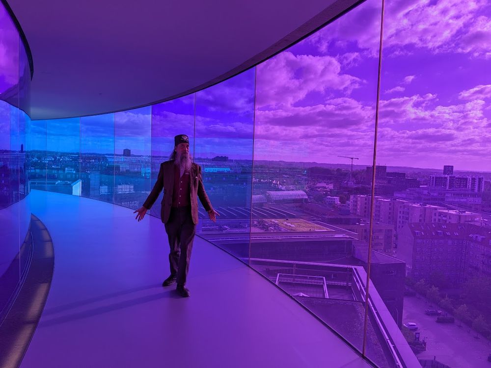 A grey-bearded man wearing a tweed jacket and a tweed fez made my Fez-O-Rama stands within the circular rooftop walkway of structural glass above the Aarhus Art Museum in Denmark. The man stands within the blue-violet spectrum area that tints the view of the entire city that color.