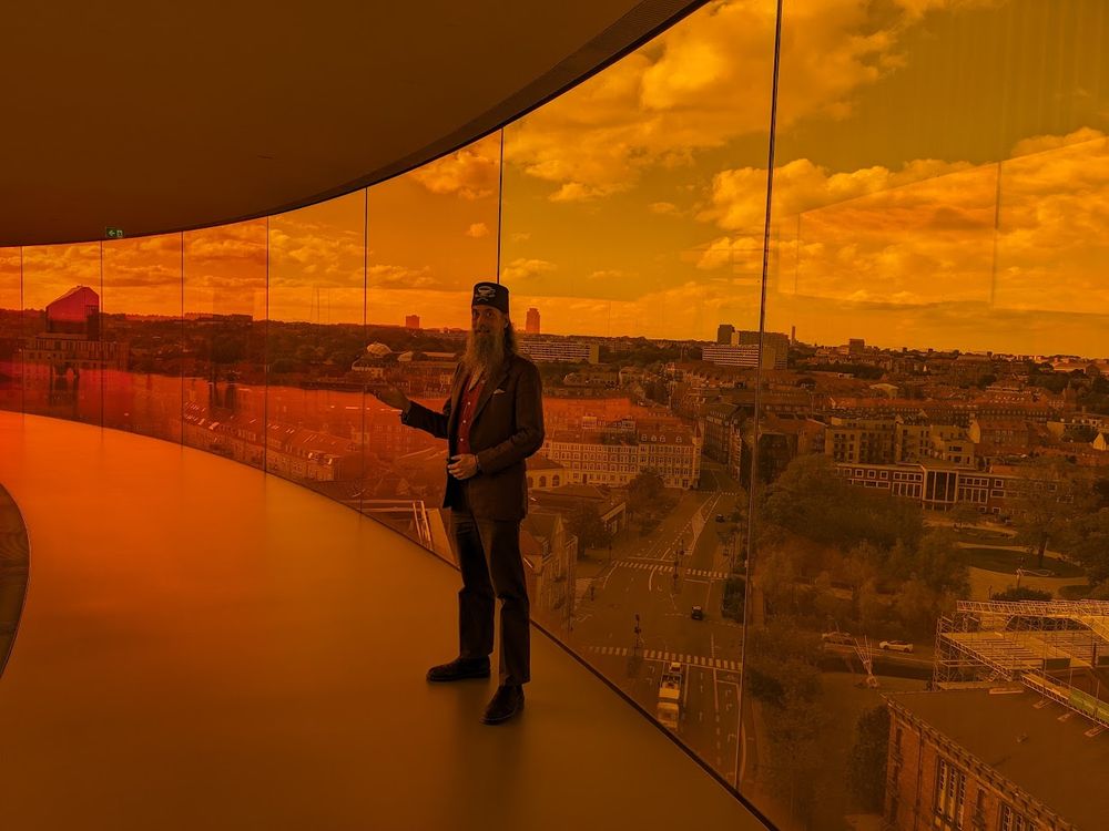 A grey-bearded man wearing a tweed jacket and a tweed fez made my Fez-O-Rama stands within the circular rooftop walkway of structural glass above the Aarhus Art Museum in Denmark. The man stands within the reddish-orange spectrum area that tints the view of the entire city that color.