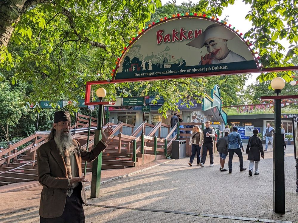 A grey-bearded man wearing a tweed jacket and a tweed fez from Fez-O-Rama motions appreciatively at an entry sign for Bakken, the world's oldest amusement park located in Denmark.