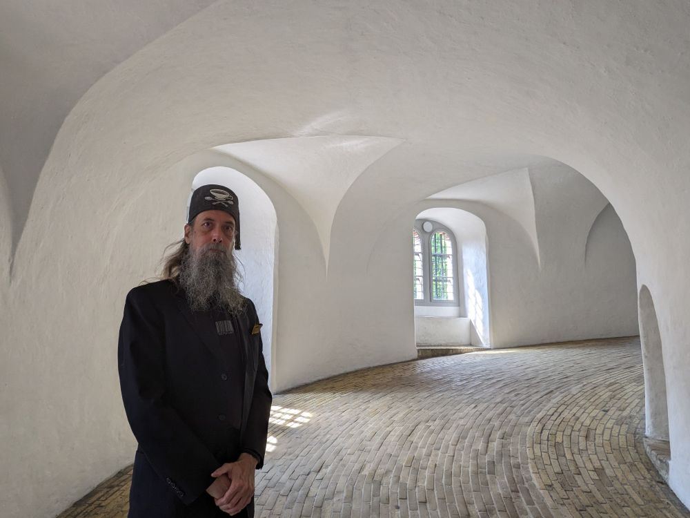 A grey-bearded man wearing a black suit and a grey tweed fez handcrafted by Fez-O-Rama stands in the interior of the Rundetaarn in Copenhagen, a cylindrical brick tower constructed in 1642. The walls are smooth white plaster transitioning into arches overhead, and the floor is a helical composition of tightly packed tan bricks.