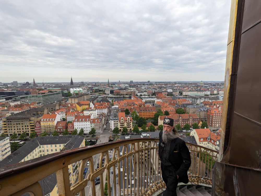 A grey-bearded man wearing a black suit and a grey tweed fez with an embroidered teacup and crossed spoons on its front fabricated by Fez-O-Rama walks up the exterior spiral staircase of Vor Frelsers Kirke, a Baroque Revival church in Copenhagen, Denmark. The point of view is higher than the overwhelming majority of the skyline of the city.