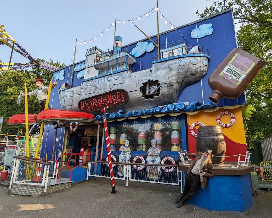A grey-bearded man wearing a tweed jacket and a tweed fez by Fez-O-Rama leans by one arm from the façade of a funhouse located in Bakken, the world's oldest amusement park located in Denmark.