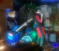 A photograph of the post-lounge musical performance group called No-Bots, taken at a basement show from their debut performance on December 31 2024.

On stage right rhythm performer Coco Nono in respendent blue dress and black jacket, wielding a blue Squier Bronco bass guitar.

On stage left vocalizer and keymaster Jojo Nono looks sharp in a black vest and red skirt, singing into a microphone. 

Behind her microNono* waits patiently upon a keyboard stand.

*microNono is an Alesis Vortex midi keytar, connected to a microKORG synthesizer and Roland SP-404.

If you wish to bathe in the sound waves of the No-Bots performance art pieces, intercept their scheduled transmission at the performance art venue Century at 1350 S 29th St, Philadelphia, PA 19146 on the evening of March 9 2025.