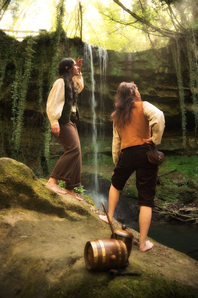 Dans un décor féérique, deux jeune hobbits femmes essaient d'observer quelque chose dans la lumière à travers les branches de la forêt. Peut être un dragon ?
Derrière elles tombe la cascade, ainsi que des lianes de lierre.
En premier plan flou, l'on peut observer la tabatière en tonneau et leurs pipes.