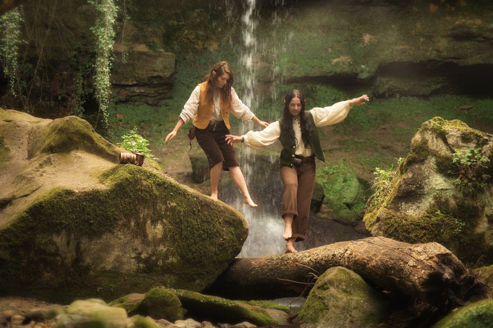 Dans un décor féérique, deux jeune hobbits femmes s'amusent à marcher sur un tronc en travers de la rivière.
Derrière elles tombe une cascade.
Elles laissent de côté sur un rocher moussu leur tabac et pipes.