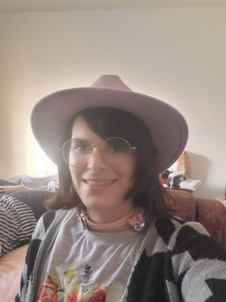 Me, a 24 year old trans woman with round glasses and neck length hair, wearing a pink hat and collar