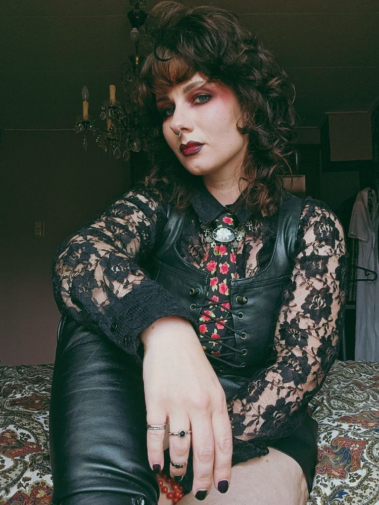 Woman in her thirties sitting on a bed, wearing black nail polish, red lipstick and eyeshadow, a black lace blouse and leather vest, and black tie with red roses. One leg is pulled up showing part of leather overknee boots. Her hair is worn in a curled wolfcut, visible jewelry are silver and black rings and brooch worn below the tie's knot.