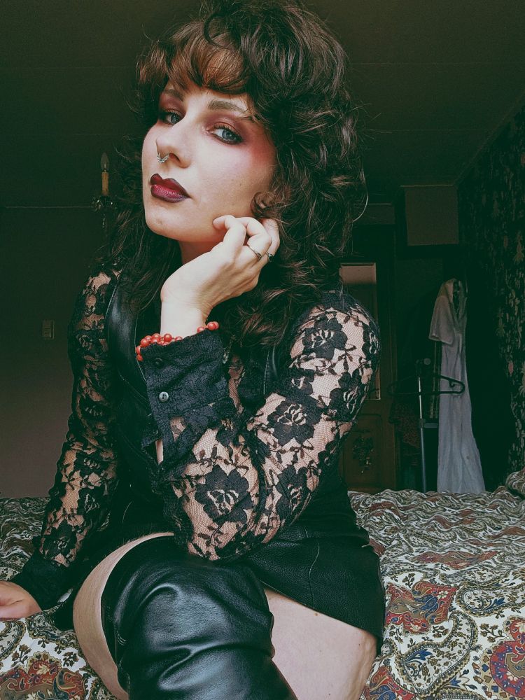 Woman in her thirties sitting on a bed, wearing black nail polish, red lipstick and eyeshadow, a black lace blouse and leather vest, and black tie with red roses. One leg is pulled up showing part of leather overknee boots. Her hair is worn in a curled wolfcut, visible jewelry are silver and black rings and a red beaded bracelet.