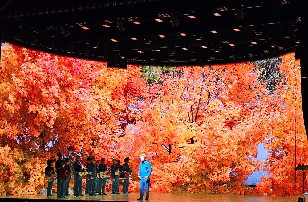 David Byrne and his backup singers and band on stage with a backdrop of vibrant orange leaves. He took this photo in Minneapolis while riding bikes with his band and crew.
